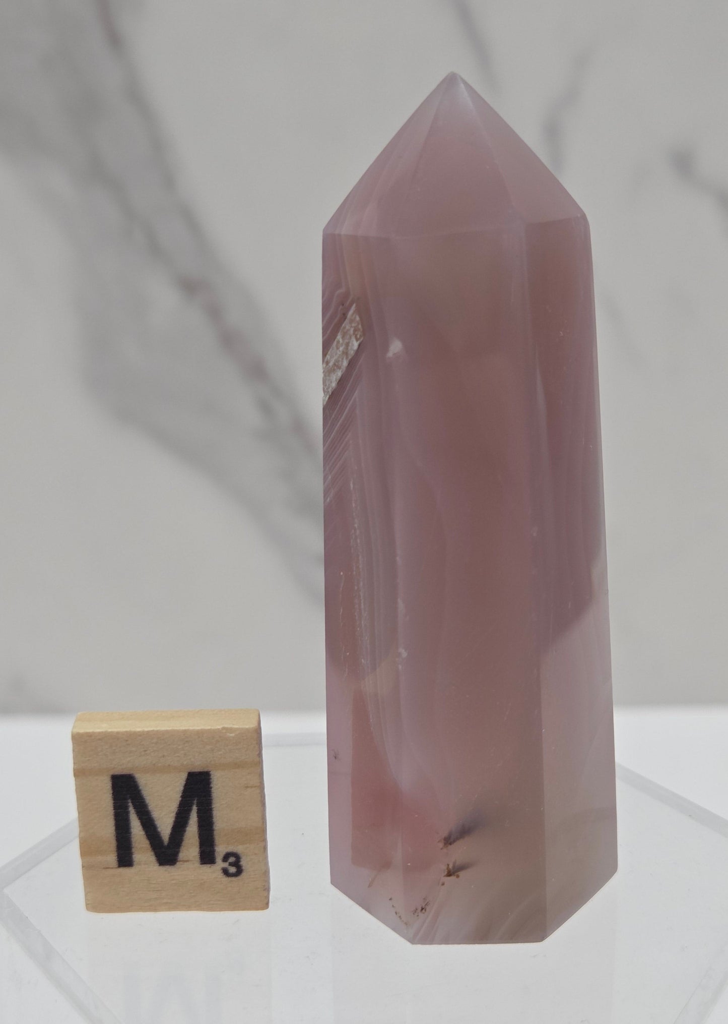 A faceted pink quartz crystal point displayed horizontally on a clear stand, with a Scrabble tile spelling 'M' nearby on a white marble surface.