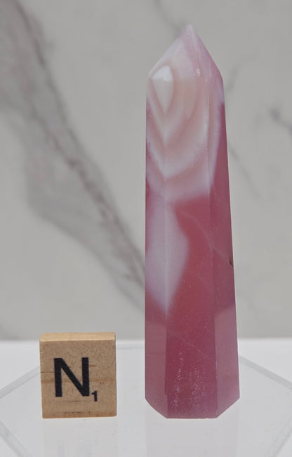 A pink and white banded quartz crystal point displayed on a clear acrylic stand next to a wooden Scrabble tile with the letter 'N', set against a white and gray marble background.
