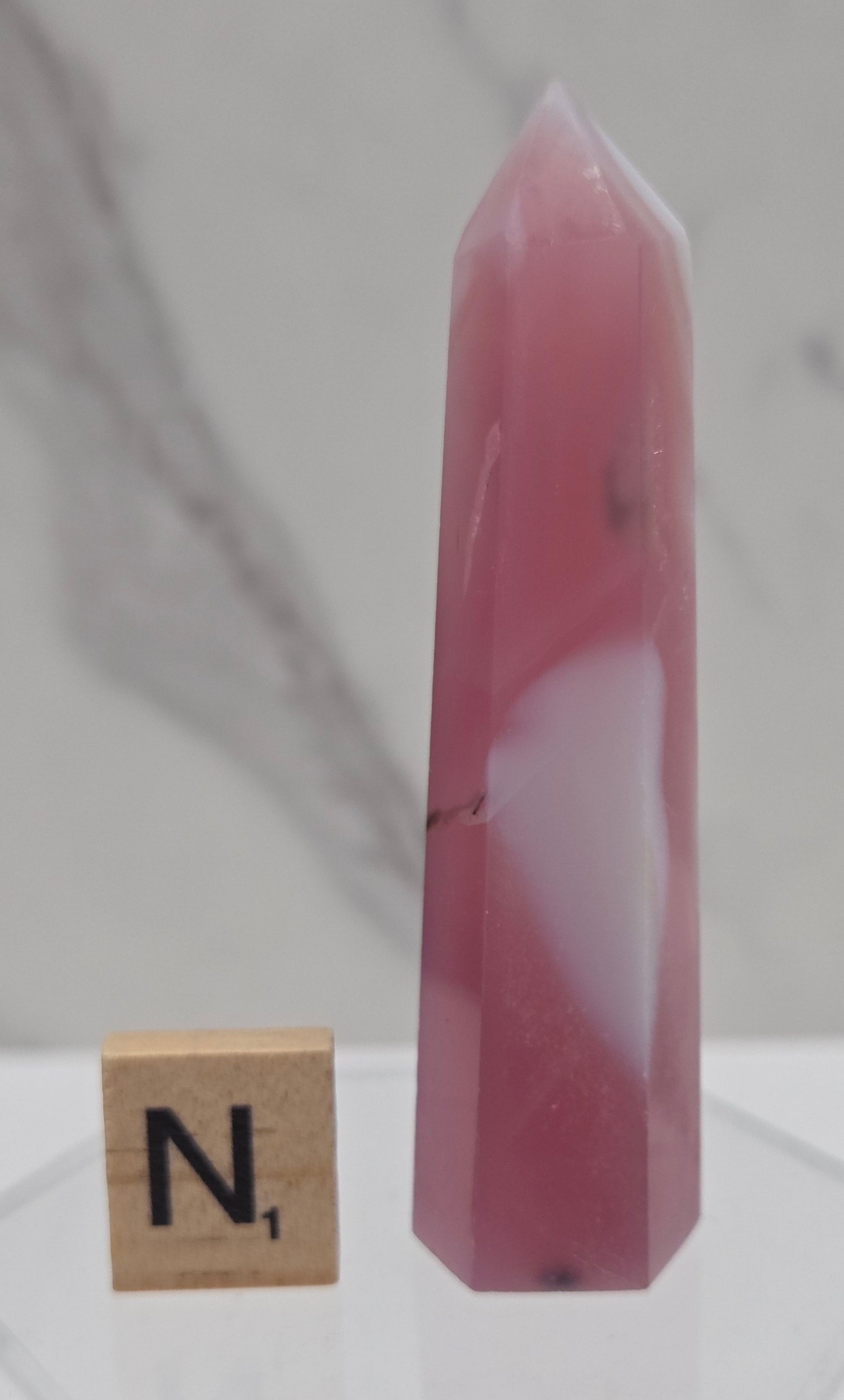 A faceted pink rose quartz crystal point displayed on a clear acrylic stand, with a wooden Scrabble tile showing the letter 'N' nearby, set against a light gray marble-patterned background.