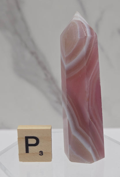 A polished pink and white agate crystal point displayed on a clear acrylic stand next to a wooden Scrabble tile with the letter P.