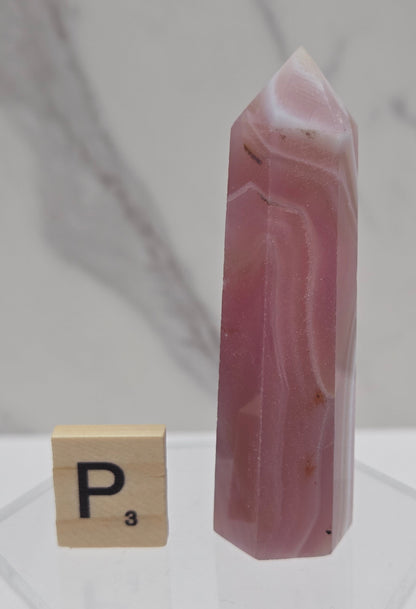 A polished pink agate crystal point with natural white and darker pink veining, displayed horizontally next to a small Scrabble tile labeled 'P' on a clear acrylic stand against a white marble-patterned background.