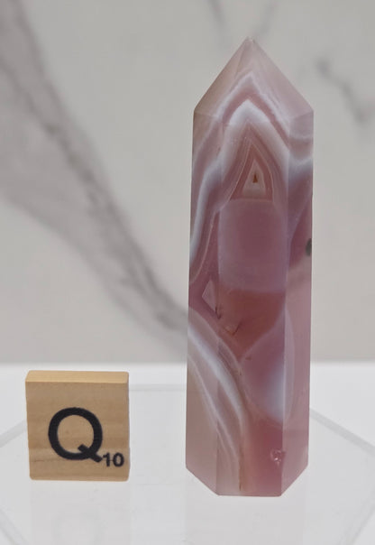 A polished pinkish-white agate crystal point with natural banding, shown floating above a small wooden Scrabble tile on a clear acrylic stand against a white marble-patterned background.