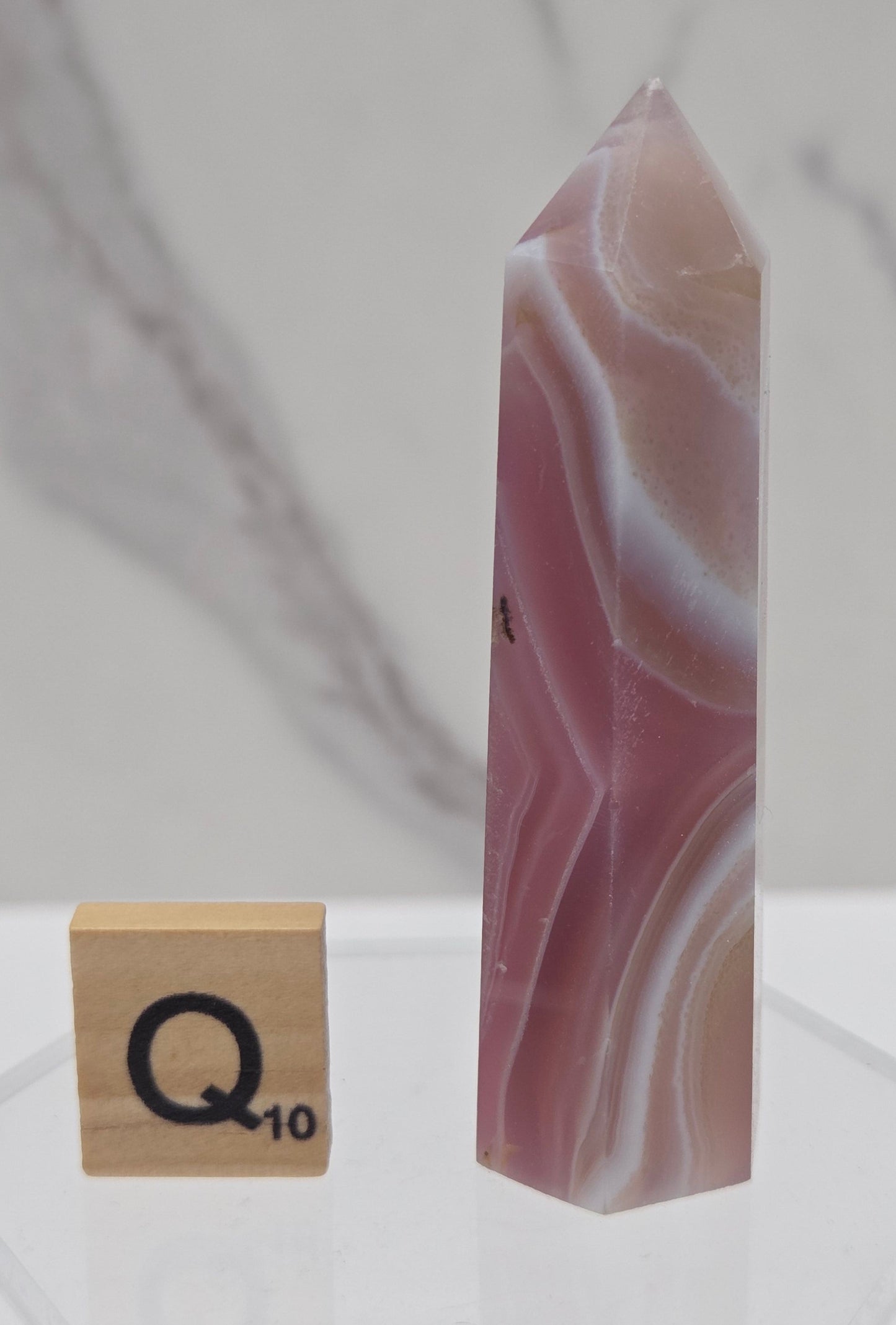 A polished pink and white agate crystal point with natural banding, displayed horizontally on a clear acrylic stand next to a wooden tile with the letter Q.