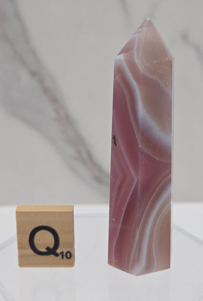 A polished pink and white agate crystal point with natural banding, displayed horizontally on a clear acrylic stand next to a wooden tile with the letter Q.