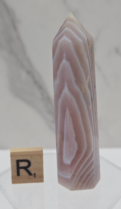 A polished rectangular agate crystal point with natural banded patterns in pale pink, beige, and white, displayed on a clear stand next to a small wooden block with a black letter 'R' on a light marble surface.