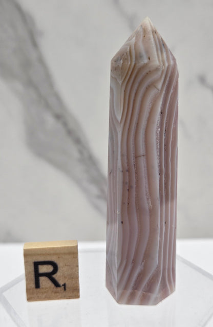 A polished agate crystal point with natural banding in beige, brown, and gray tones, displayed horizontally on a clear acrylic stand next to a wooden block with the letter R.