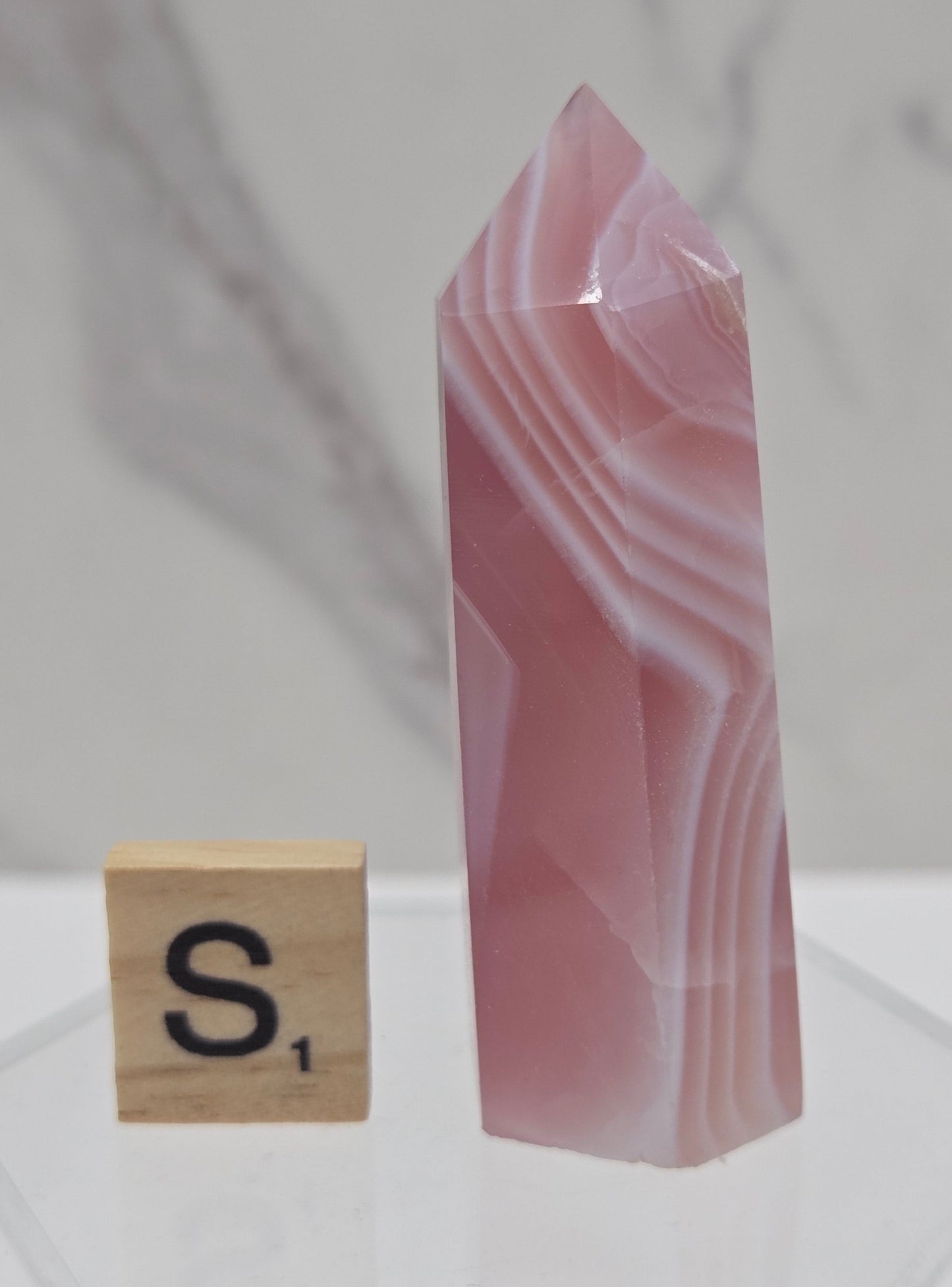 A pink and white banded agate crystal point displayed horizontally on a clear acrylic stand, with a wooden Scrabble tile showing the letter 'S' placed below it, against a light gray marble-patterned background.