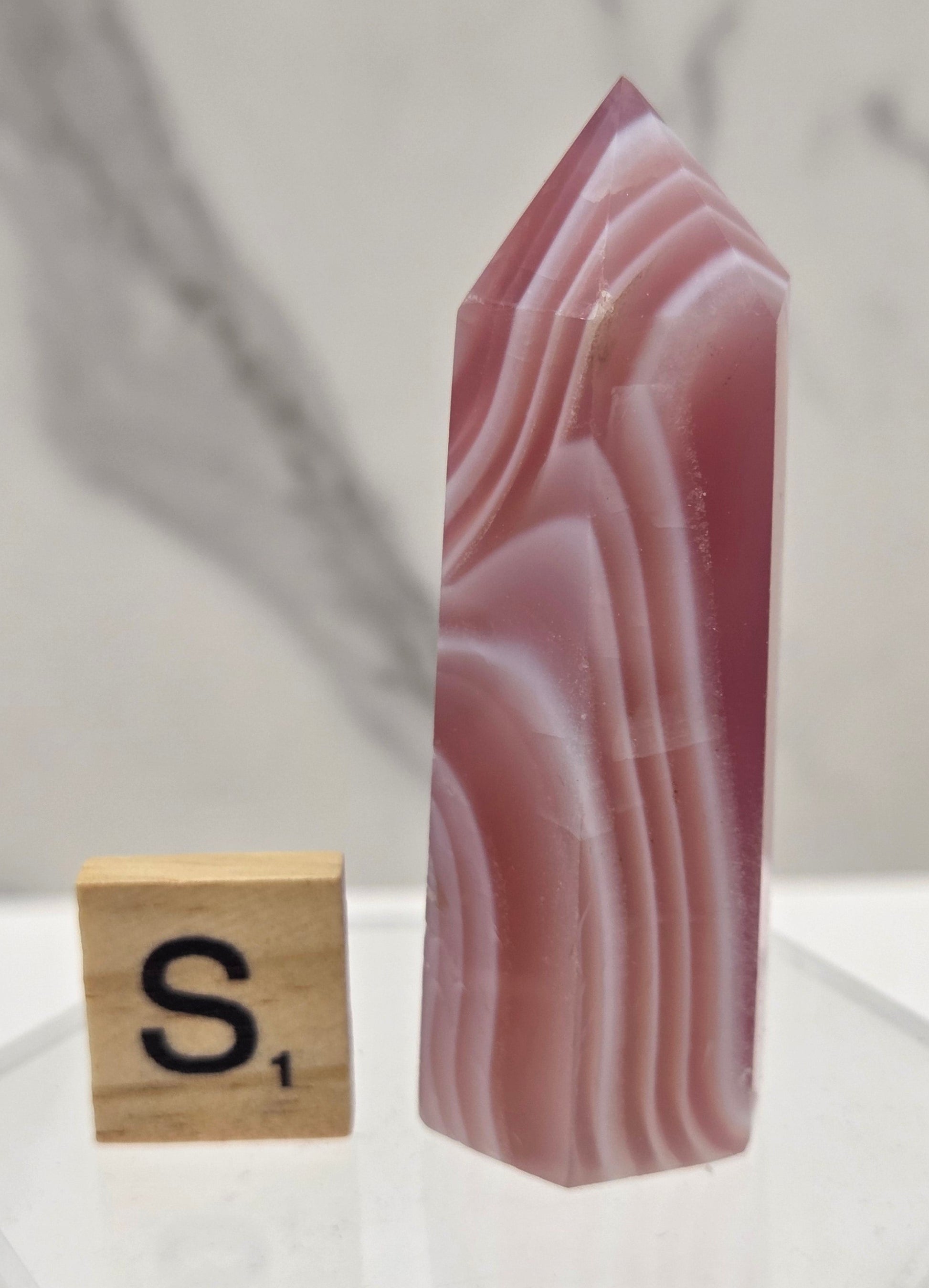 A polished pink agate crystal point with natural banding, displayed on a clear acrylic stand next to a small wooden block with a black 'S' symbol, against a light gray marble background.