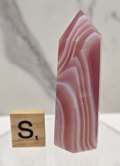 A polished pink agate crystal point with natural banding, displayed on a clear acrylic stand next to a small wooden block with a black 'S' symbol, against a light gray marble background.