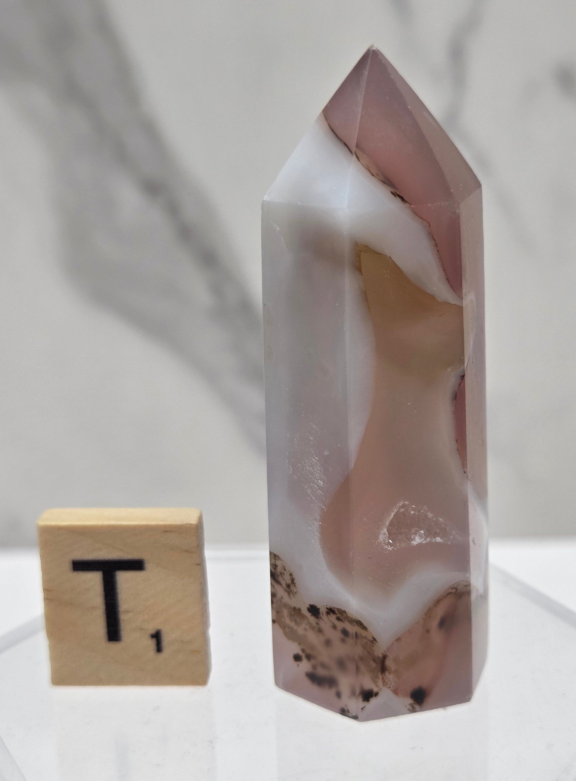 A polished agate crystal point with pink, white, and beige natural banding, displayed on a clear acrylic stand next to a wooden Scrabble tile on a white marble surface.