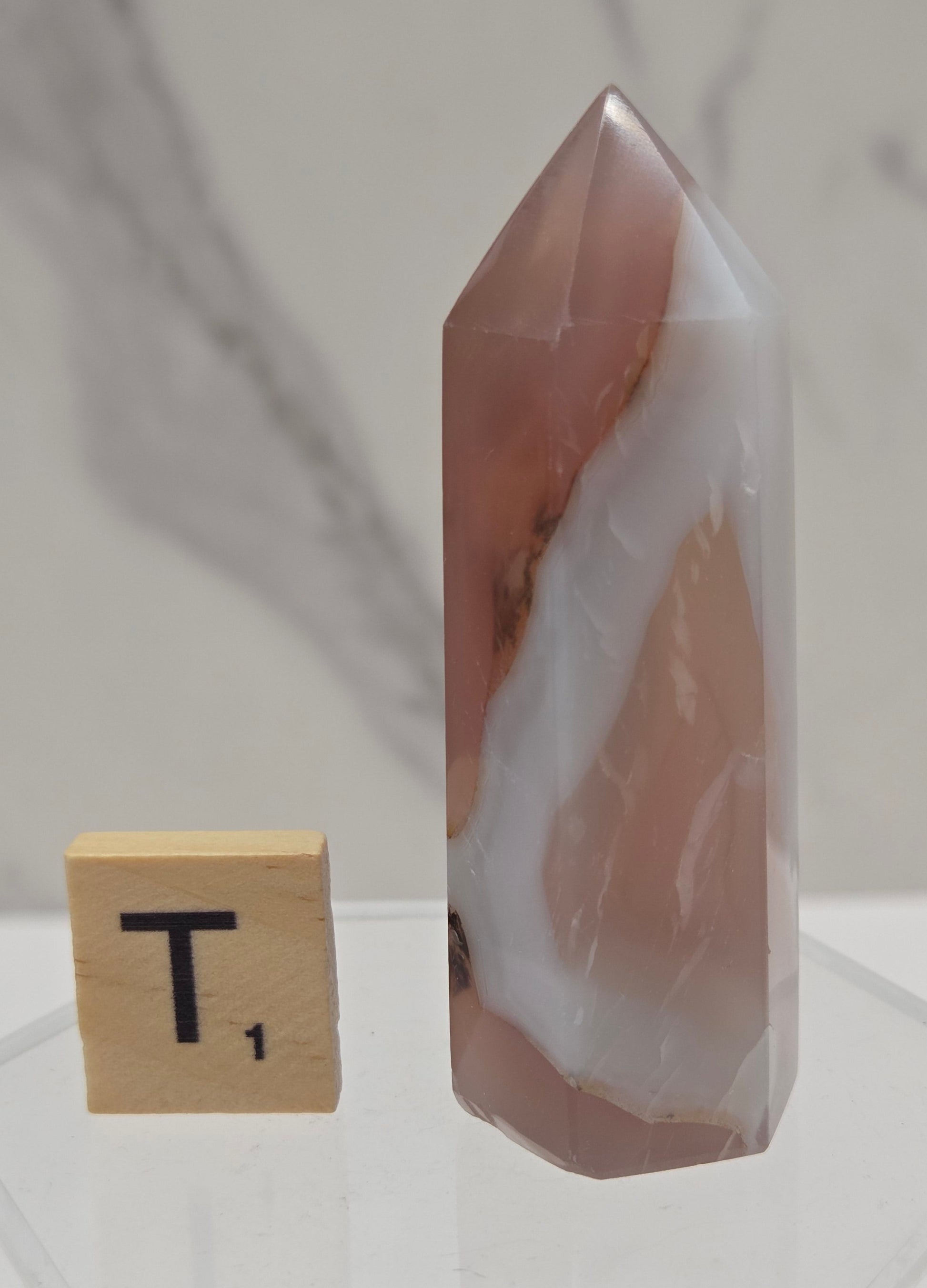 A polished rose quartz crystal point with natural white and pinkish-brown banding, displayed on a clear acrylic stand next to a small wooden Scrabble tile on a light marble surface.