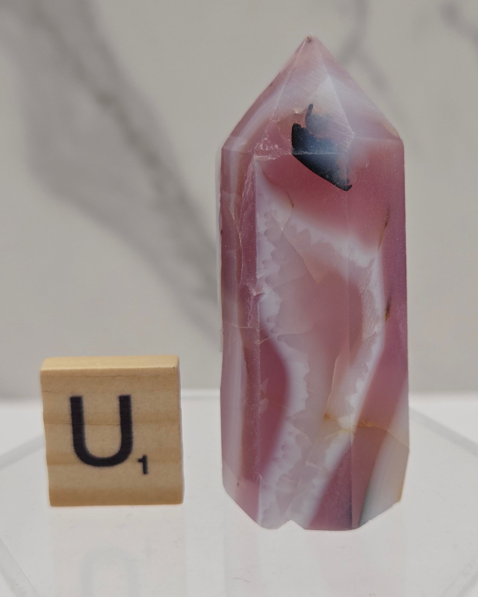 A pink and white agate crystal point with a dark inclusions, displayed on a clear acrylic stand next to a wooden letter tile with the letter 'U'.