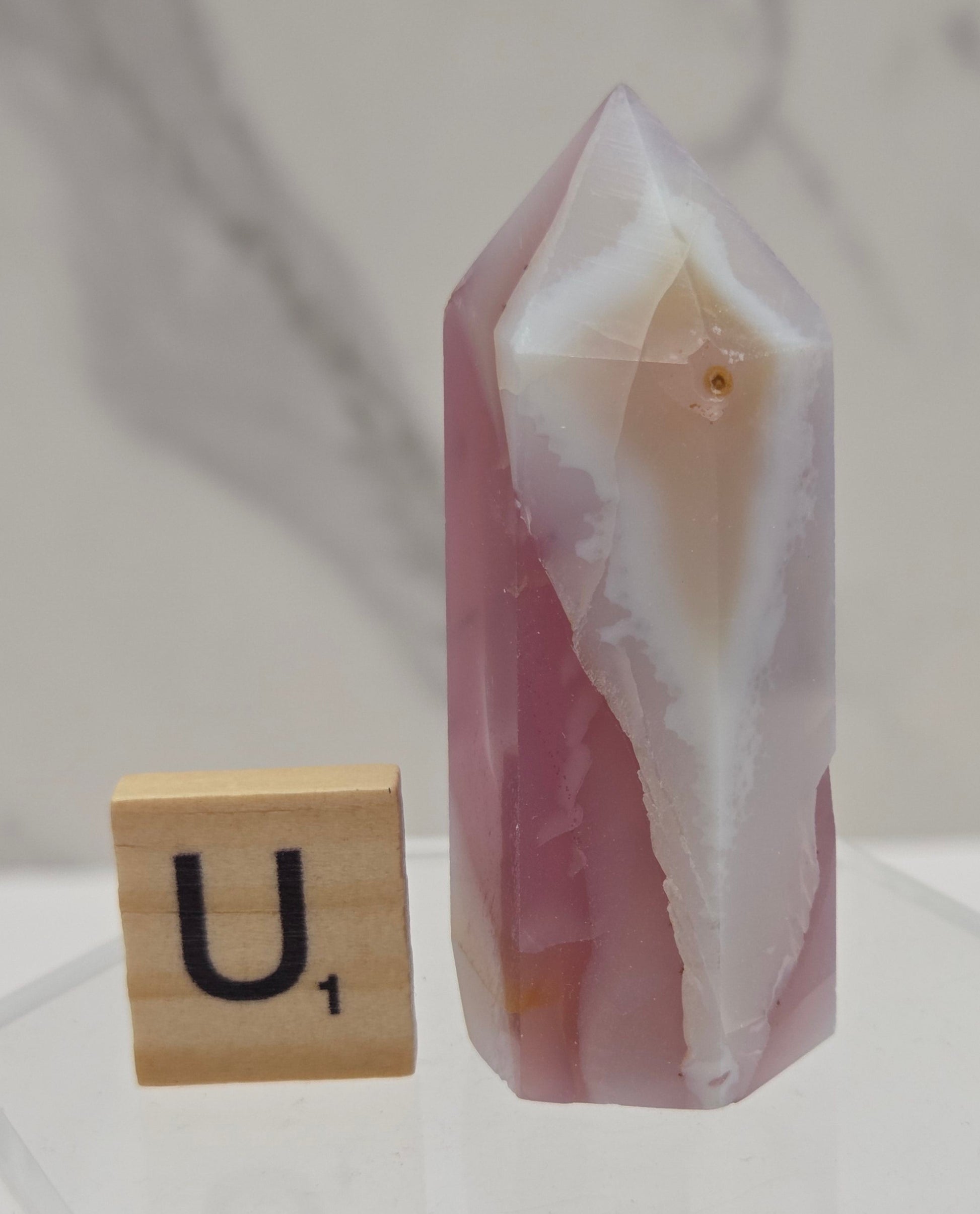 A polished pink and white agate crystal point displayed horizontally on a clear acrylic stand, with a small wooden letter tile showing the letter 'U' placed below it.
