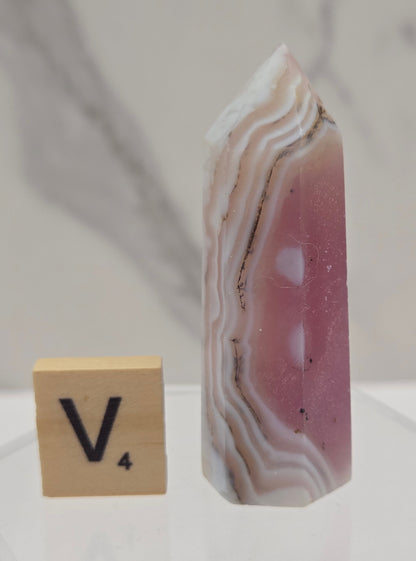 A polished pink and white agate crystal point with natural banded layers, displayed next to a wooden Scrabble tile on a clear acrylic stand against a light marble background.