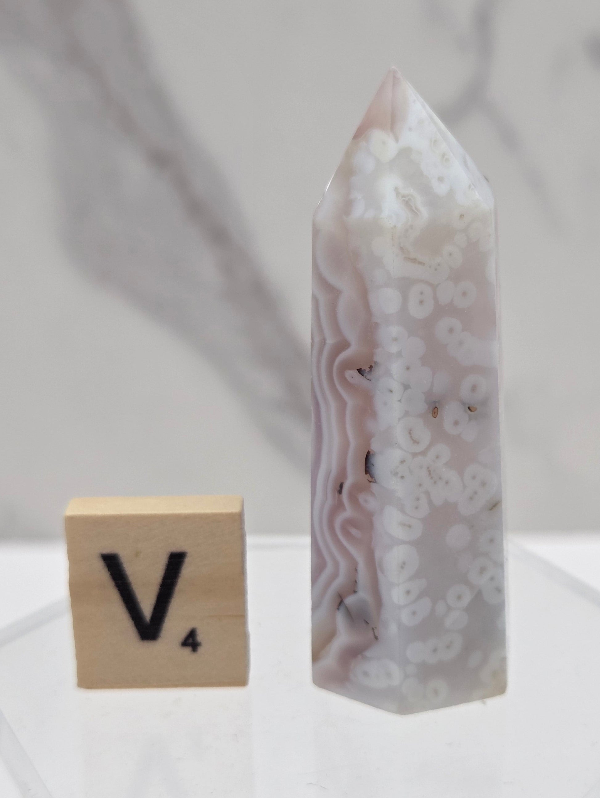 A polished agate crystal point with natural banded patterns in white, gray, and soft pink, displayed next to a wooden tile with the letter V on a clear acrylic stand.