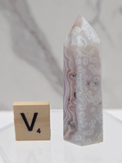 A polished agate crystal point with natural banded patterns in white, gray, and soft pink, displayed next to a wooden tile with the letter V on a clear acrylic stand.
