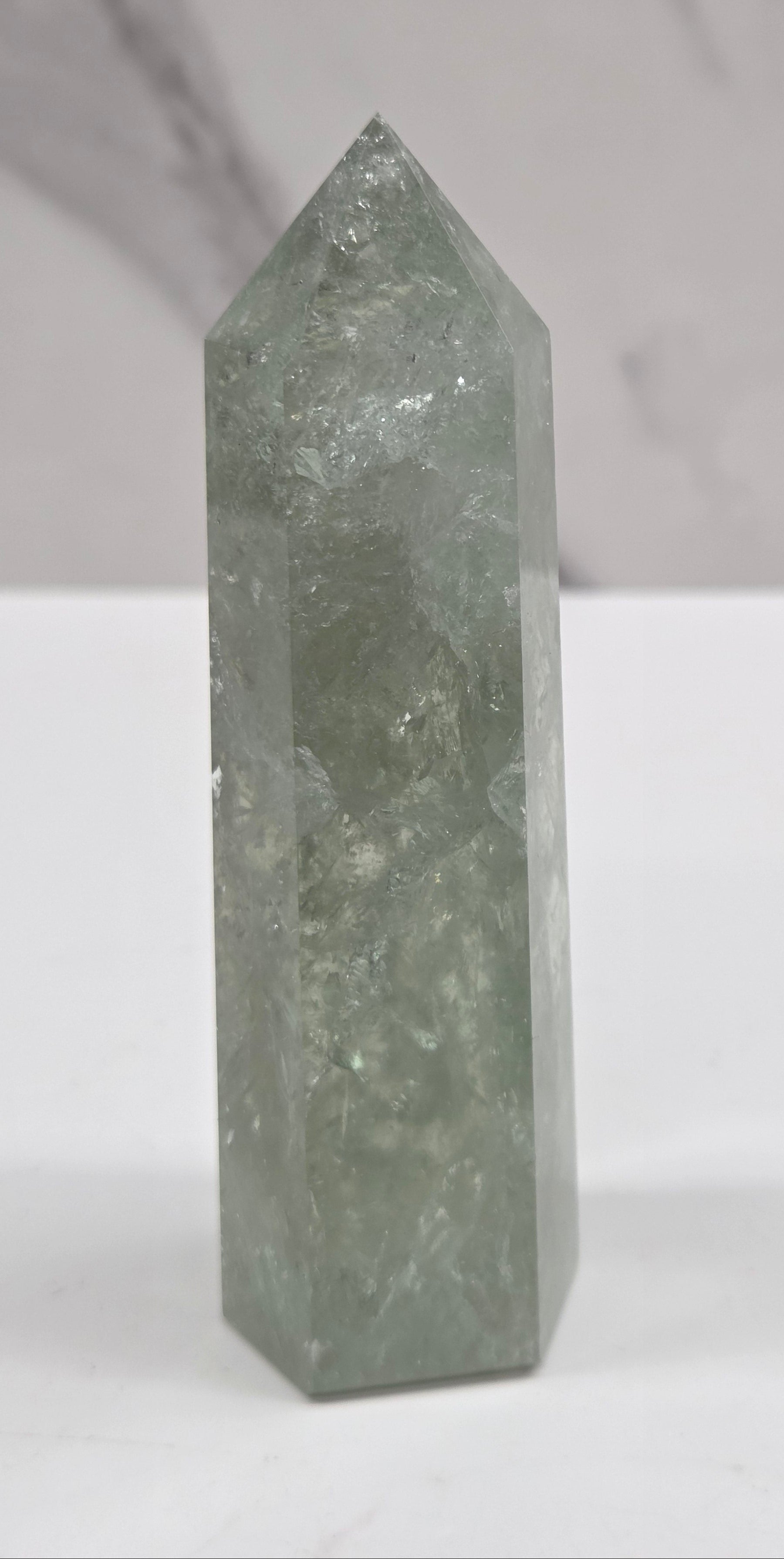 A polished green crystal point displayed horizontally against a split white and light gray marble background.