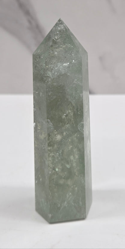 A polished green crystal point displayed horizontally against a split white and light gray marble background.