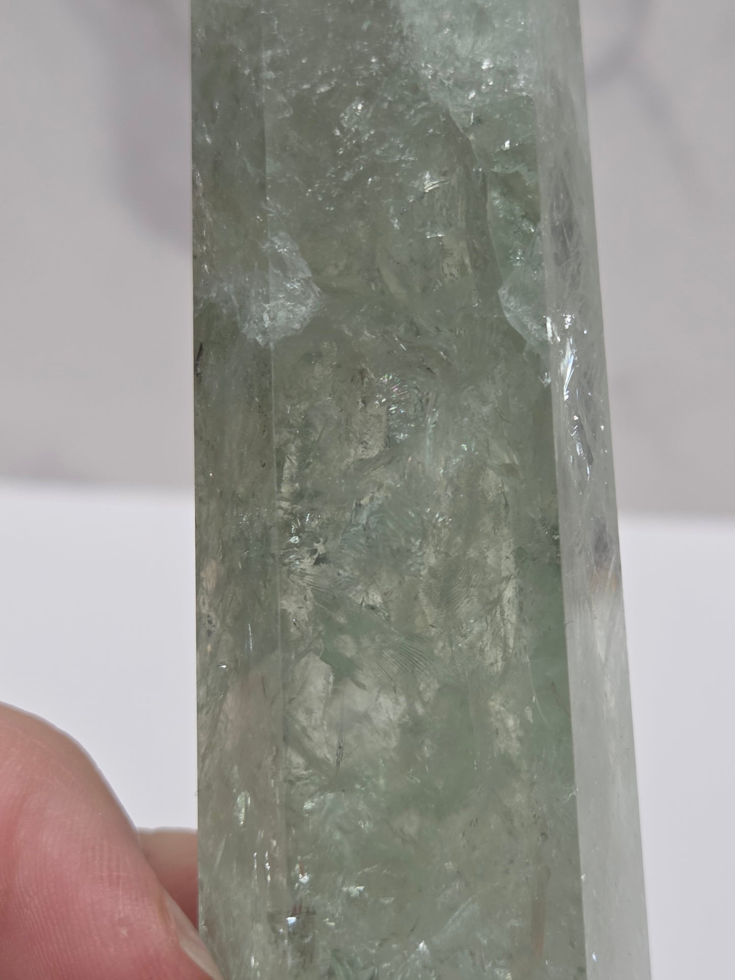 A close-up of a translucent pale green crystal or mineral specimen held by a person's fingers against a light neutral background.