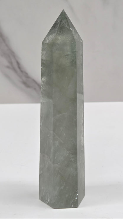 A polished gray crystal point wand or omen stone shown horizontally against a white background with a marble-textured section on the left.