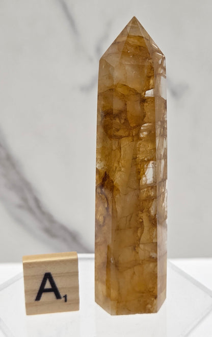 A natural golden-brown quartz crystal point displayed horizontally on a clear acrylic stand, with a small Scrabble tile showing the letter A- placed below it, set against a white marble-patterned background.
