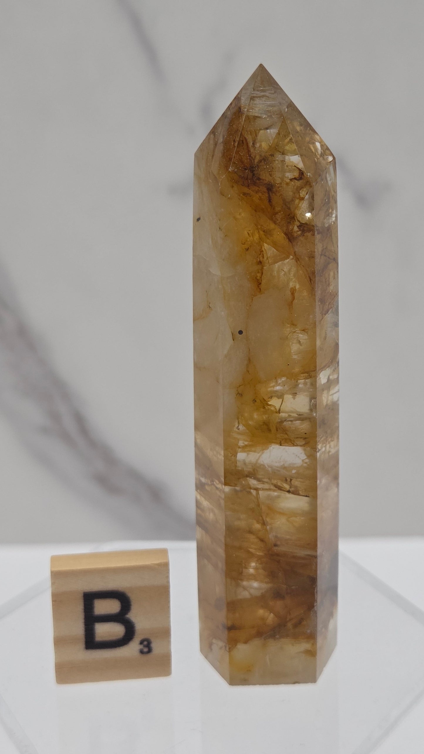 A natural quartz crystal point with golden-brown and translucent amber tones, displayed on a white marble surface next to a wooden block with the letter B.