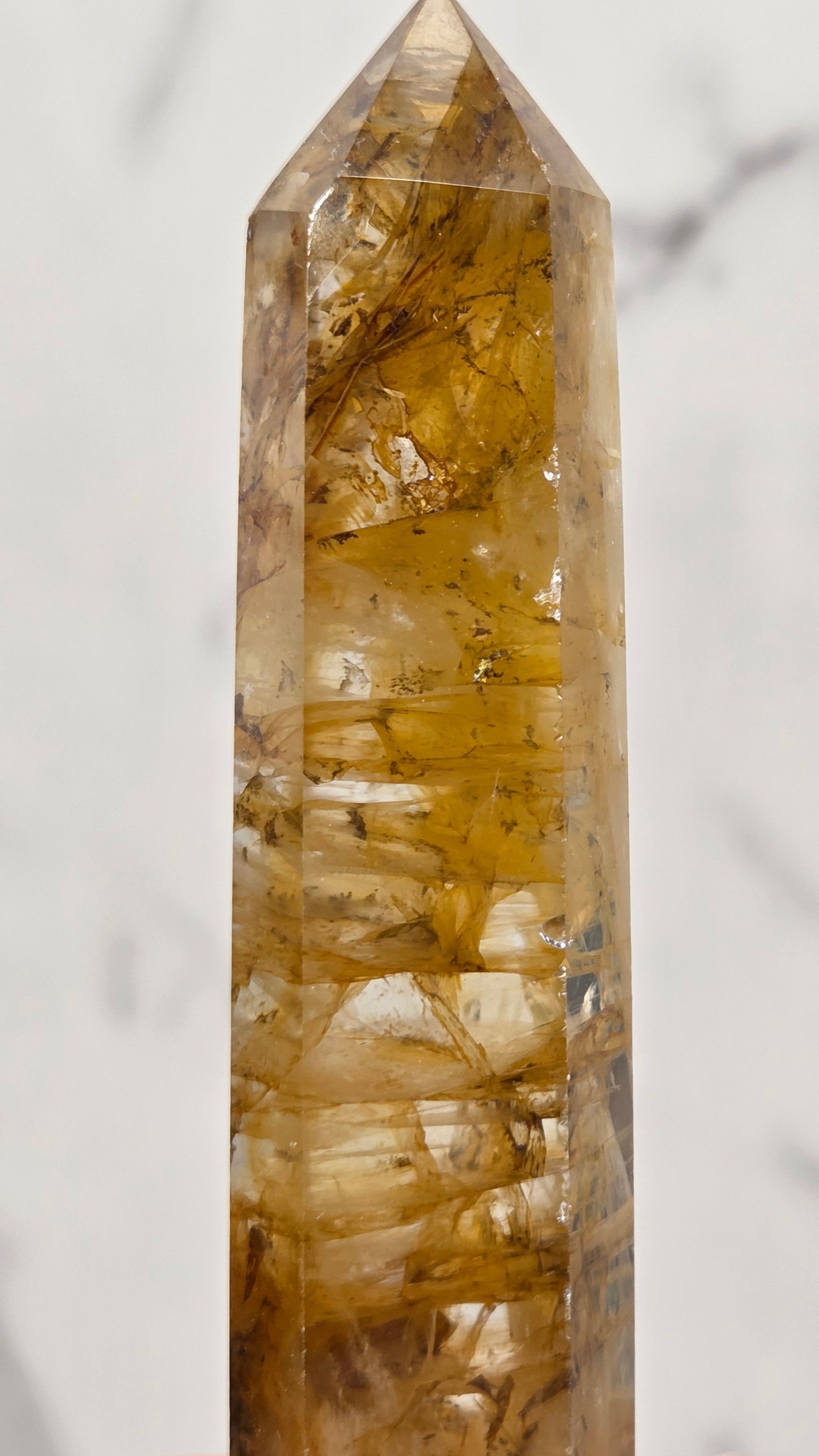 A pointed clear quartz crystal tower with prominent golden-brown and amber inclusions, shown in a close-up horizontal shot against a light gray marble background.