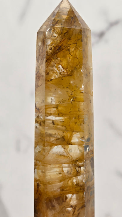 A pointed clear quartz crystal tower with prominent golden-brown and amber inclusions, shown in a close-up horizontal shot against a light gray marble background.