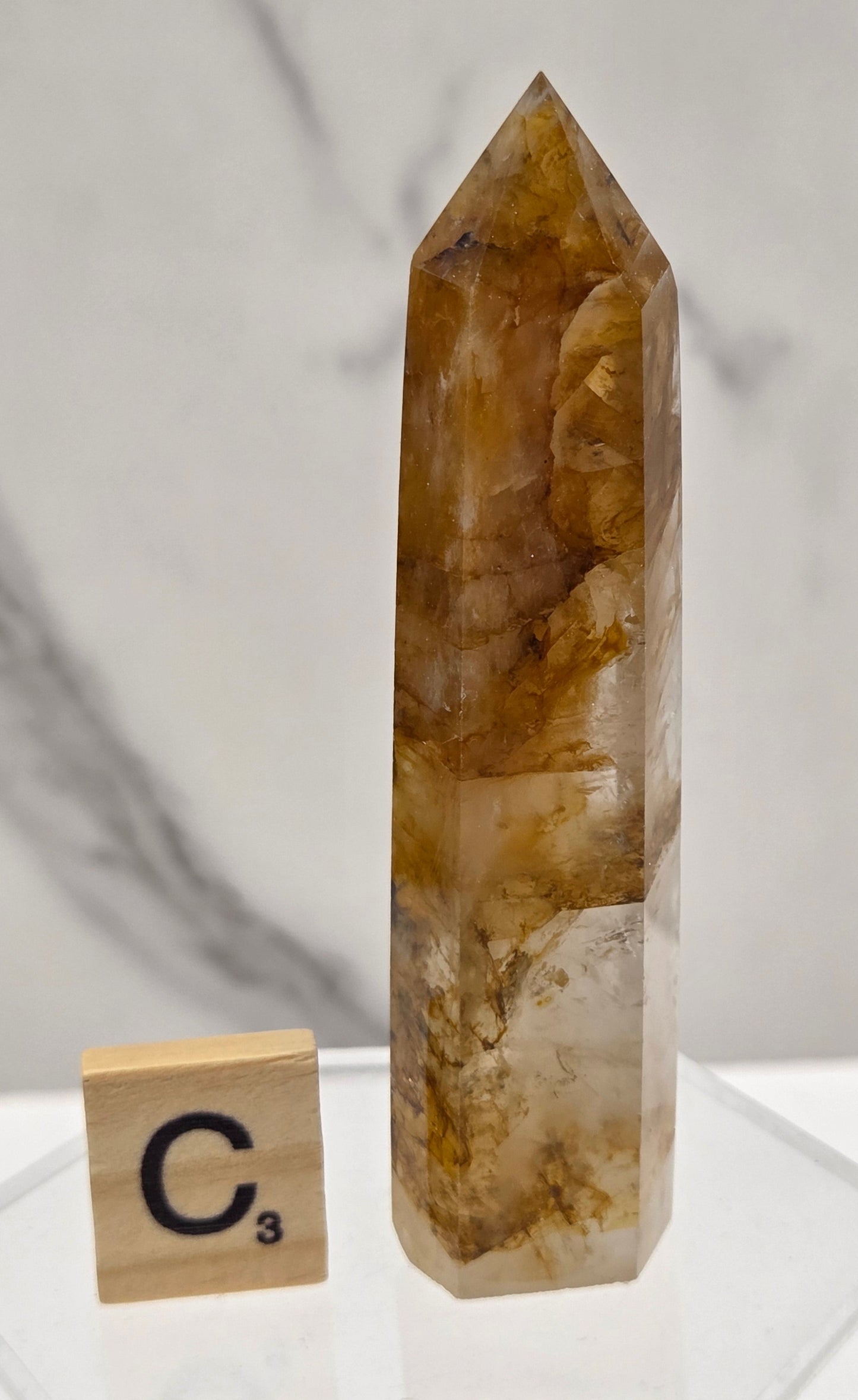 A natural quartz crystal point with brown and amber inclusions, displayed on a clear acrylic stand next to a wooden tile labeled C3 on a white marble surface.