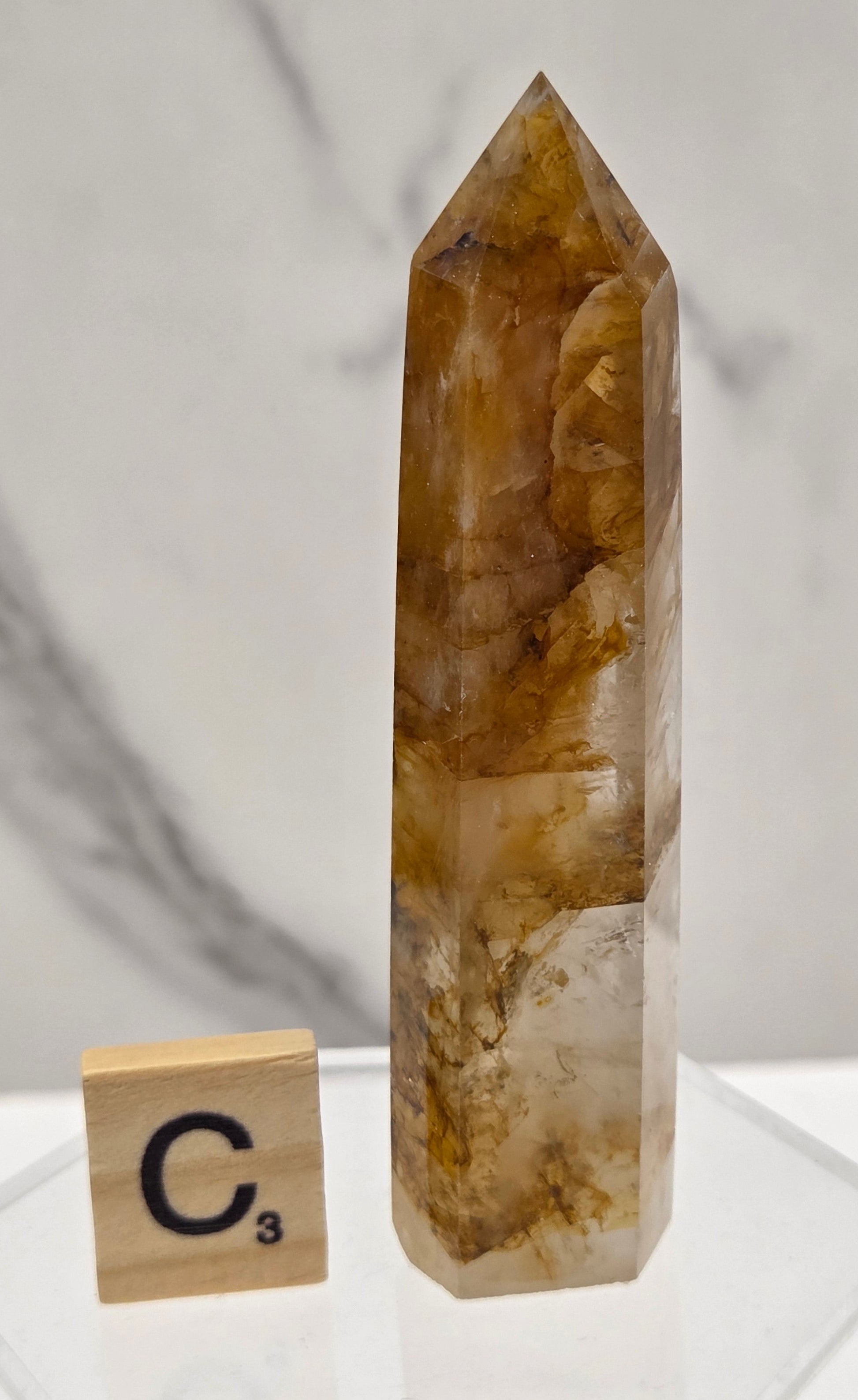 A natural quartz crystal point with brown and amber inclusions, displayed on a clear acrylic stand next to a wooden tile labeled C3 on a white marble surface.
