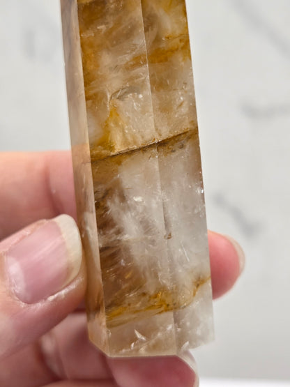 A close-up of a hand holding a faceted clear crystal point with golden-brown and white inclusions, set against a light marble-like background.