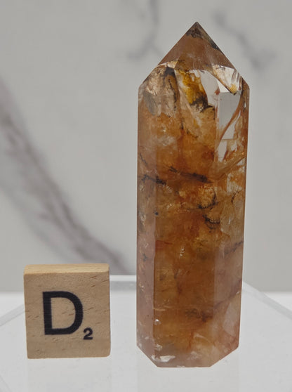 A natural clear quartz point crystal with prominent golden-brown and amber inclusions, displayed next to a wooden 'D2' tile on a light gray marble surface.