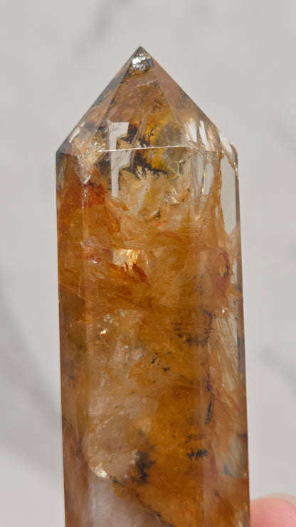 A close-up of a clear quartz crystal point with golden-brown and amber inclusions, held by a hand against a light gray marble background.