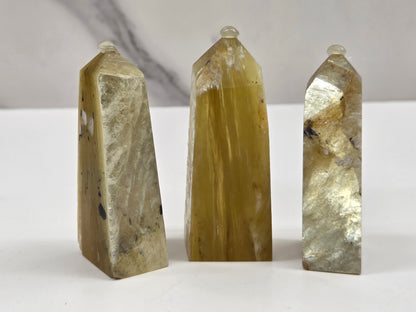 Three polished natural quartz crystal points of varying heights and sizes, showcasing golden yellow, beige, and gray marbled patterns, arranged on a white surface against a light gray marble background.