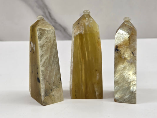 Three polished natural quartz crystal points of varying heights and sizes, showcasing golden yellow, beige, and gray marbled patterns, arranged on a white surface against a light gray marble background.