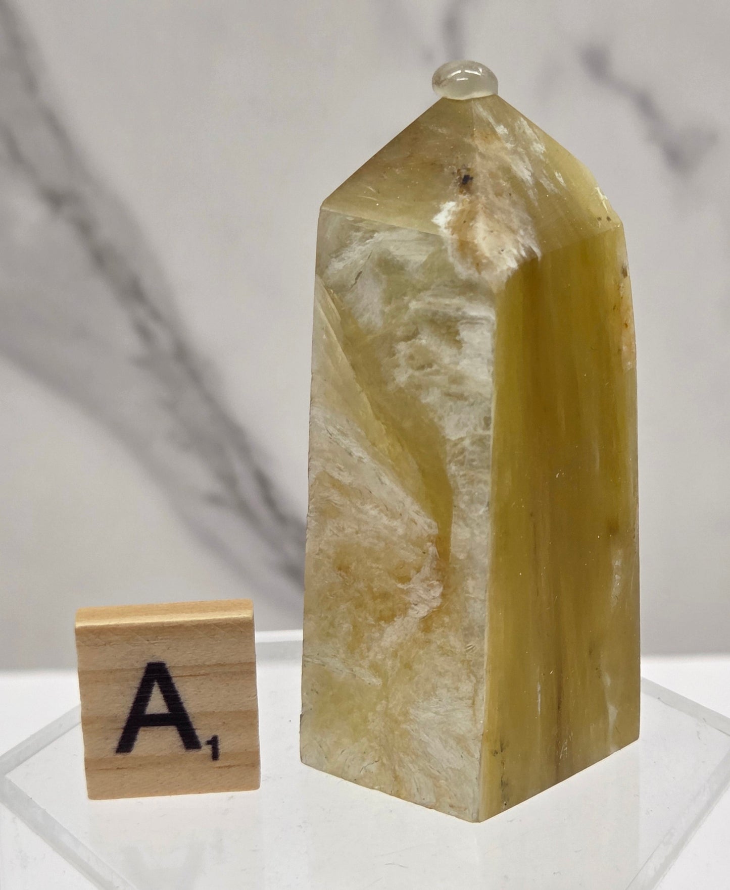 A polished yellow-green crystal point with natural striations, displayed on a clear acrylic stand next to a wooden letter tile showing the letter 'A', against a white marble background.