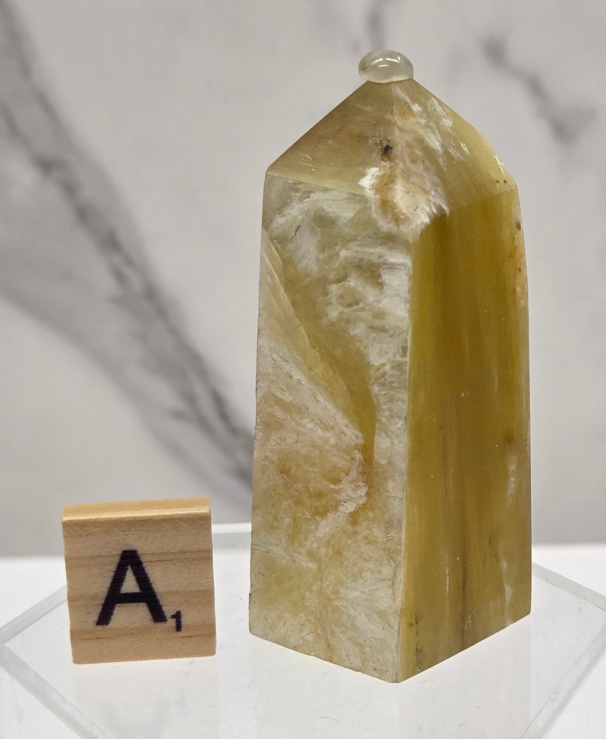 A polished yellow-green crystal point with natural striations, displayed on a clear acrylic stand next to a wooden letter tile showing the letter 'A', against a white marble background.