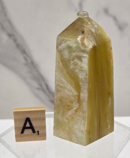 A polished yellow-green crystal point with natural striations, displayed on a clear acrylic stand next to a wooden letter tile showing the letter 'A', against a white marble background.