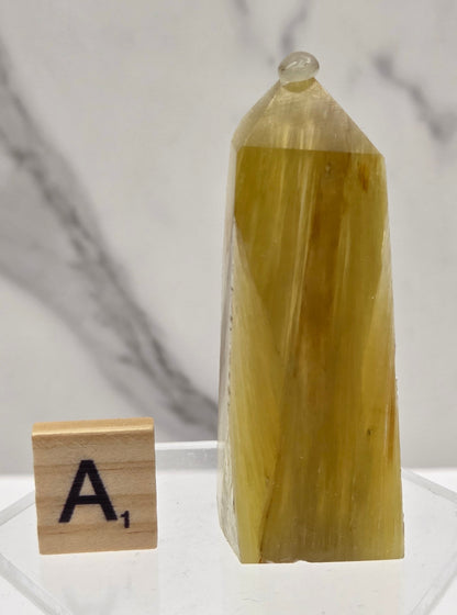 A pale yellow translucent crystal or stone specimen displayed on a clear acrylic stand, with a small Scrabble tile labeled 'A1' nearby on a white marble surface.