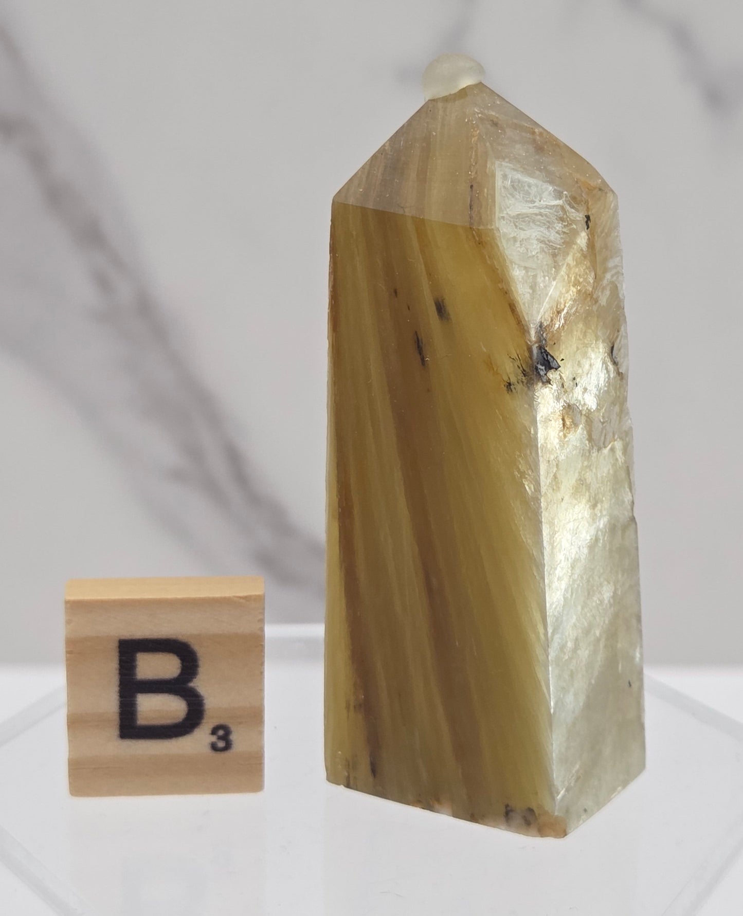 A polished golden-yellow quartz crystal point displayed on a clear acrylic stand, with a small wooden letter block showing the letter 'B' placed below it, set against a white marble-patterned background.