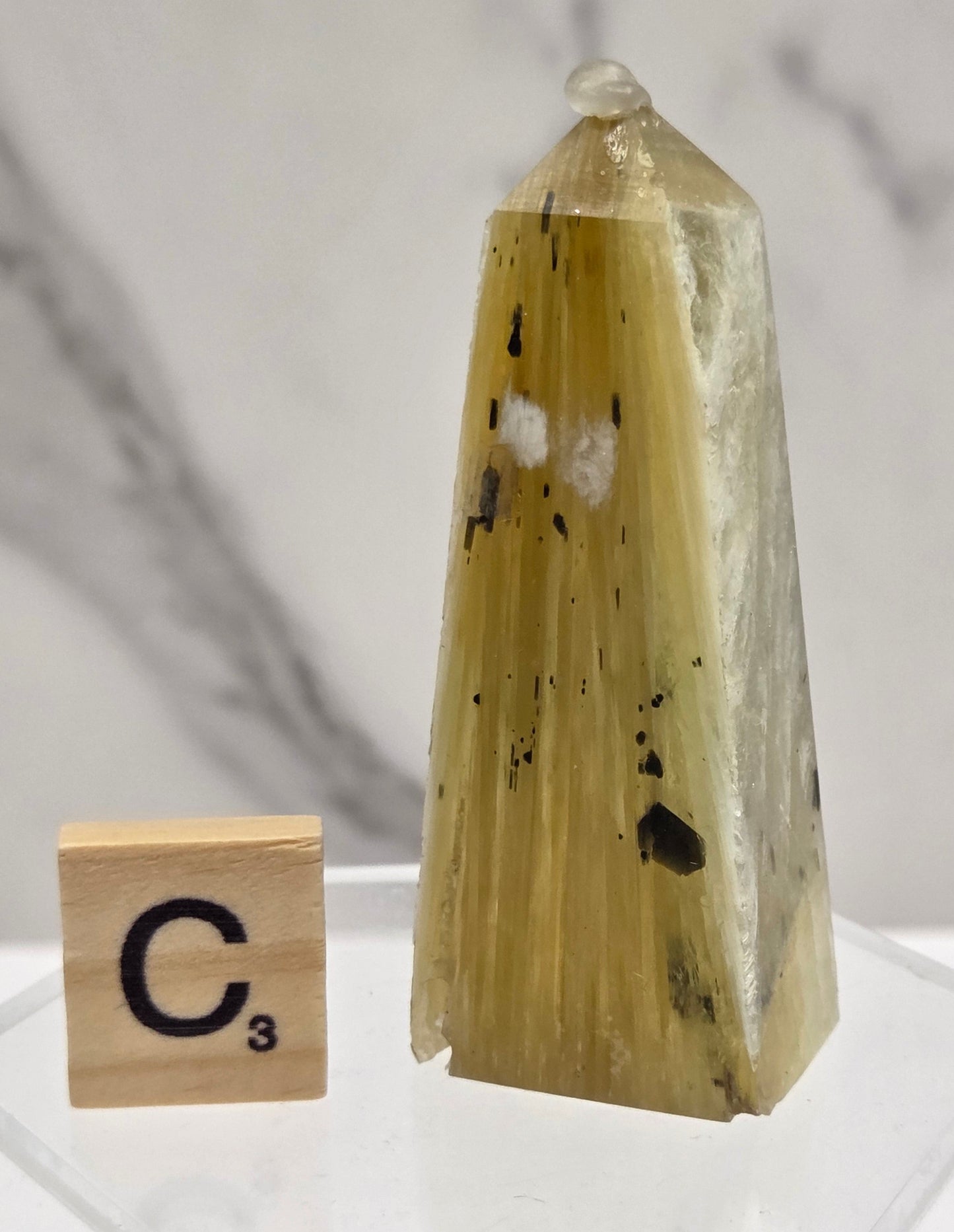 A pointed crystal specimen with a pale yellow-green translucent body and dark inclusions, displayed on a clear stand against a light marble background, with a small labeled tile nearby.