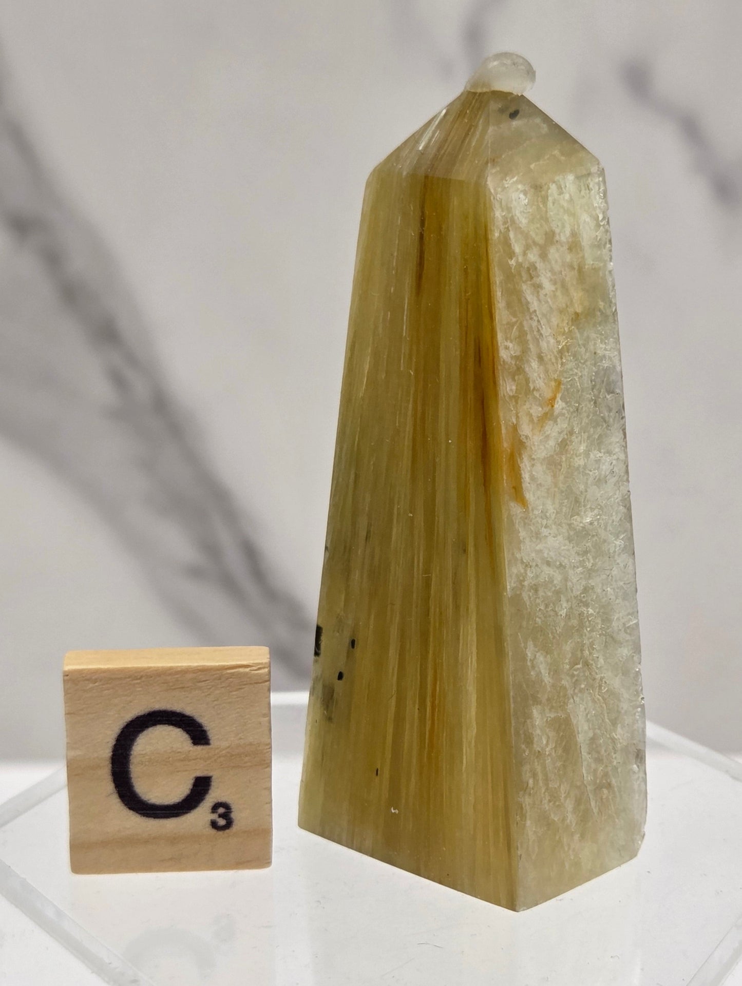 A polished yellow-green quartz point crystal displayed on a clear acrylic stand next to a wooden Scrabble tile showing the letter 'C' and value 3, set against a light marble background.
