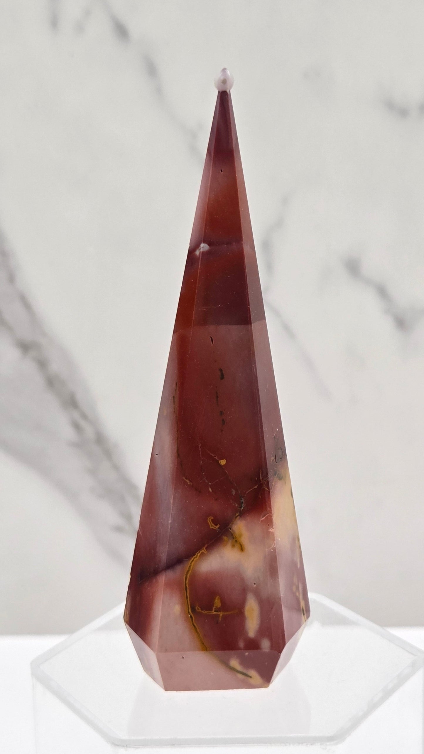 A faceted crystal point in a reddish-brown hue with natural inclusions, mounted on a clear acrylic stand against a white marble background.