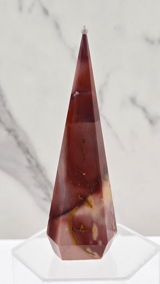 A faceted crystal point in a reddish-brown hue with natural inclusions, mounted on a clear acrylic stand against a white marble background.