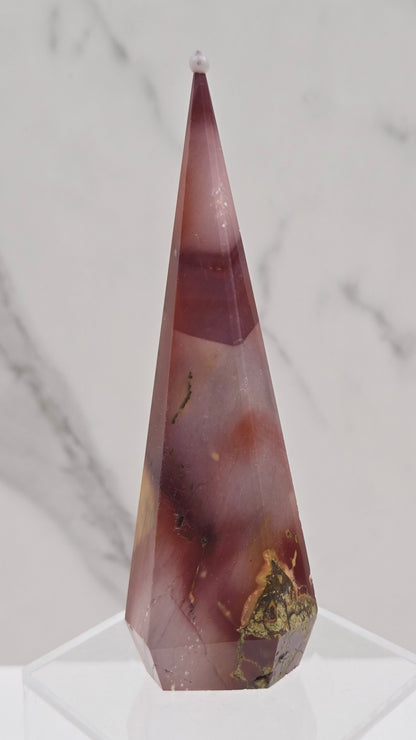 A faceted crystal point tower in a dusty rose and burgundy hue with natural inclusions, mounted on a clear acrylic display stand against a white marble background.
