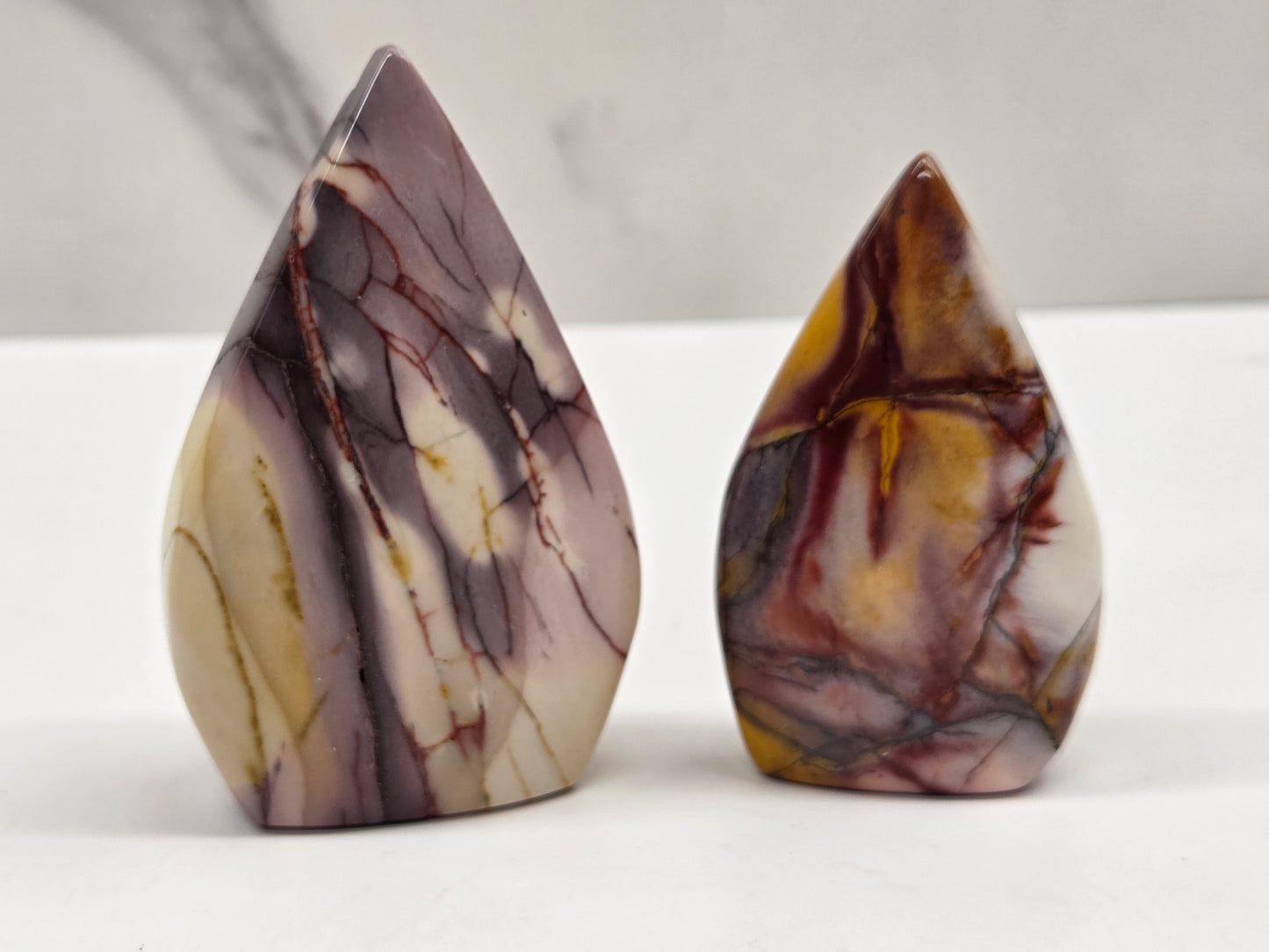 Two polished teardrop-shaped stone carvings with natural marbled patterns, one in cream and brown with dark veins and the other in brown, gold, and gray on a white surface.