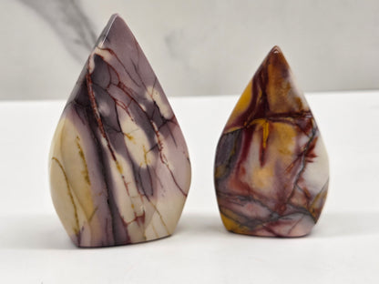 Two polished teardrop-shaped stone carvings with natural marbled patterns, one in cream and brown with dark veins and the other in brown, gold, and gray on a white surface.