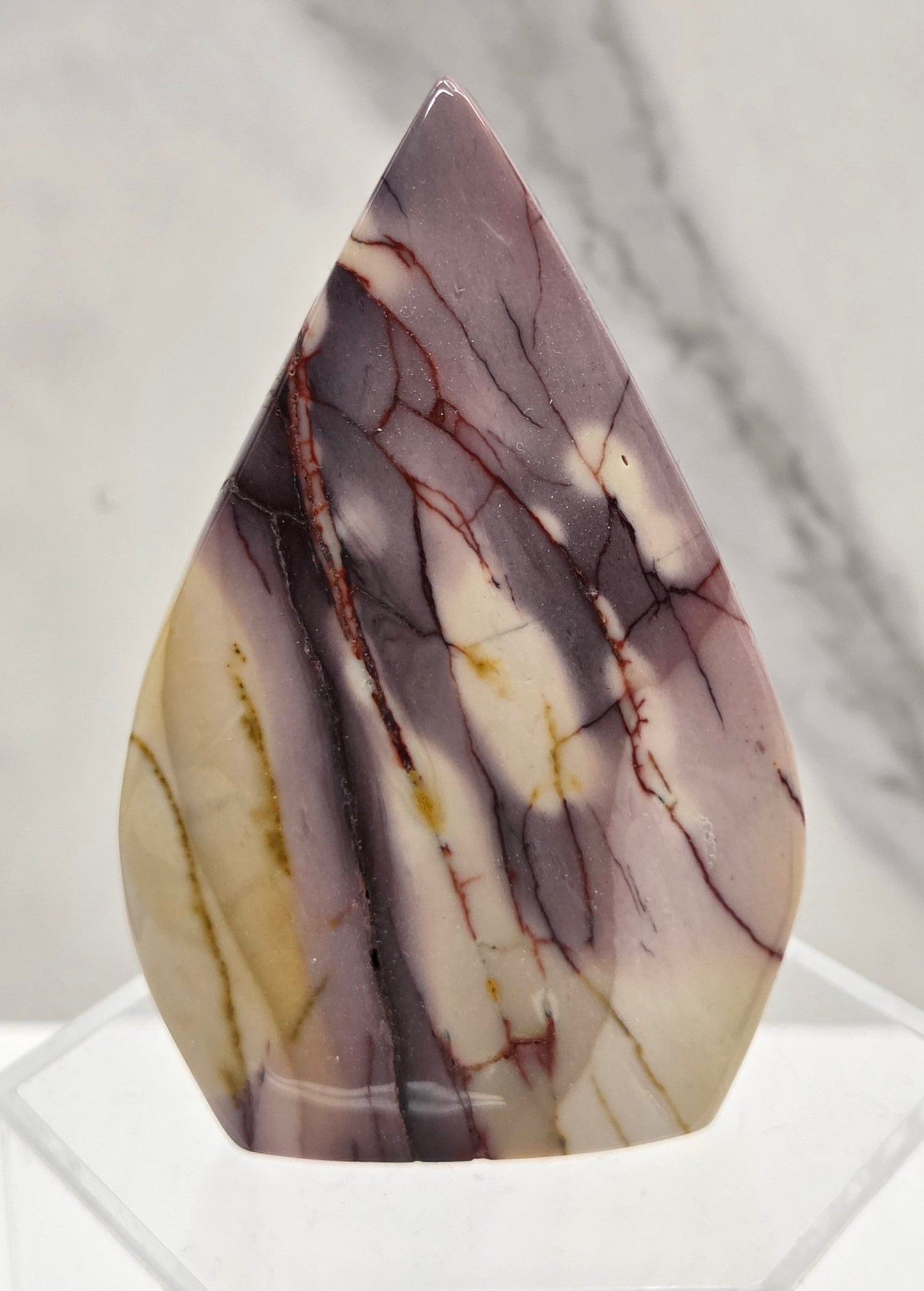 A polished teardrop-shaped cabochon gemstone with a marbled pattern of purple, cream, and brown veins, displayed on a clear acrylic stand against a light gray marble background.