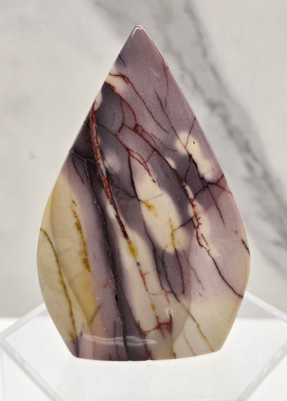 A polished teardrop-shaped cabochon gemstone with a marbled pattern of purple, cream, and brown veins, displayed on a clear acrylic stand against a light gray marble background.