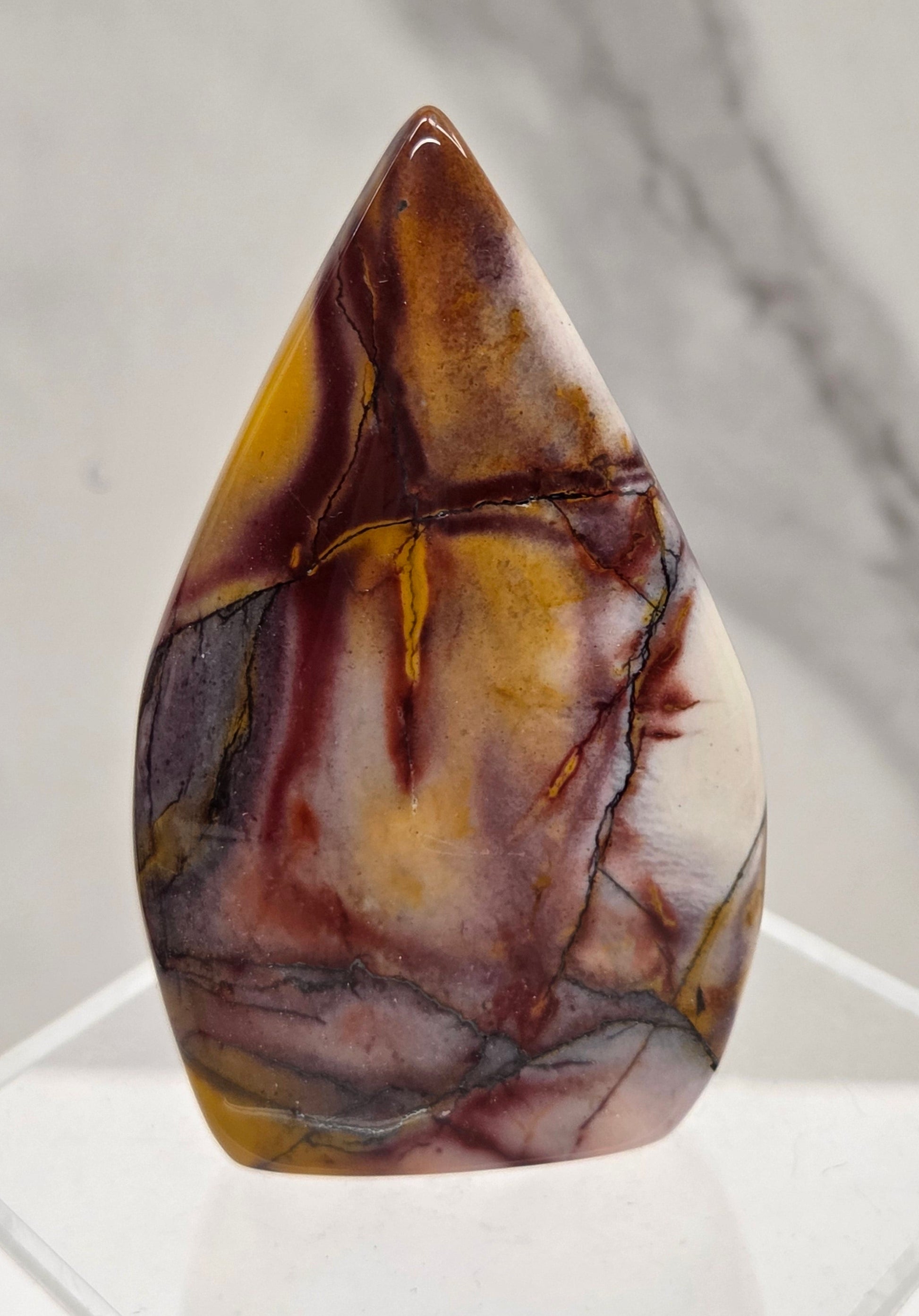 A polished teardrop-shaped cabochon gemstone with marbled brown, gold, and cream patterns, displayed on a clear acrylic stand against a light gray marble background.